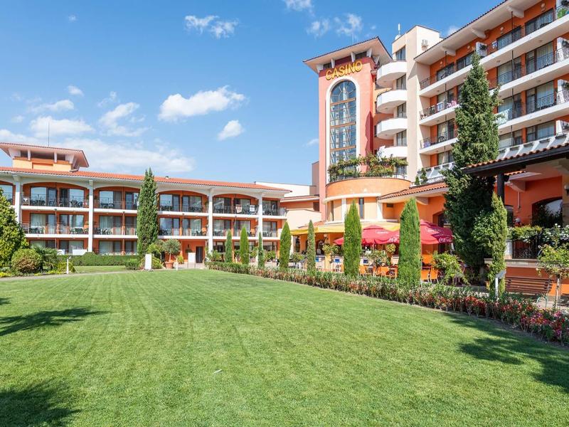 Hotel with large lawn, multi-story building, and sunny outdoor area with trees and chairs.