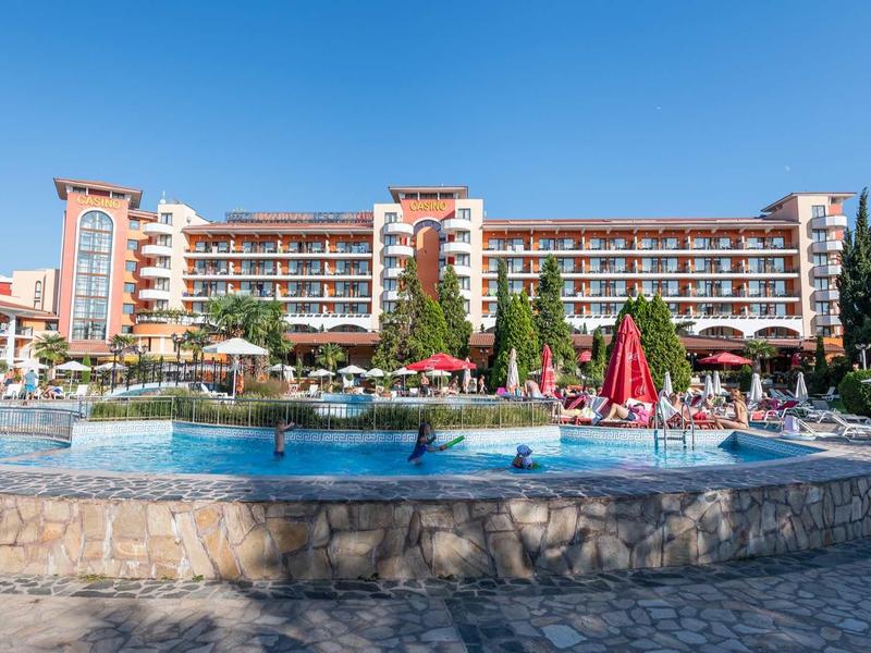 Large hotel with pool, sun umbrellas, and lounge chairs under a bright blue sky.