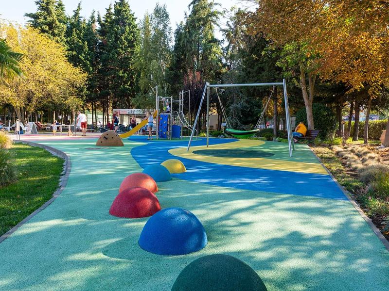 Colorful playground with swings, climbing equipment, and rubber mounds in a park with autumn trees.