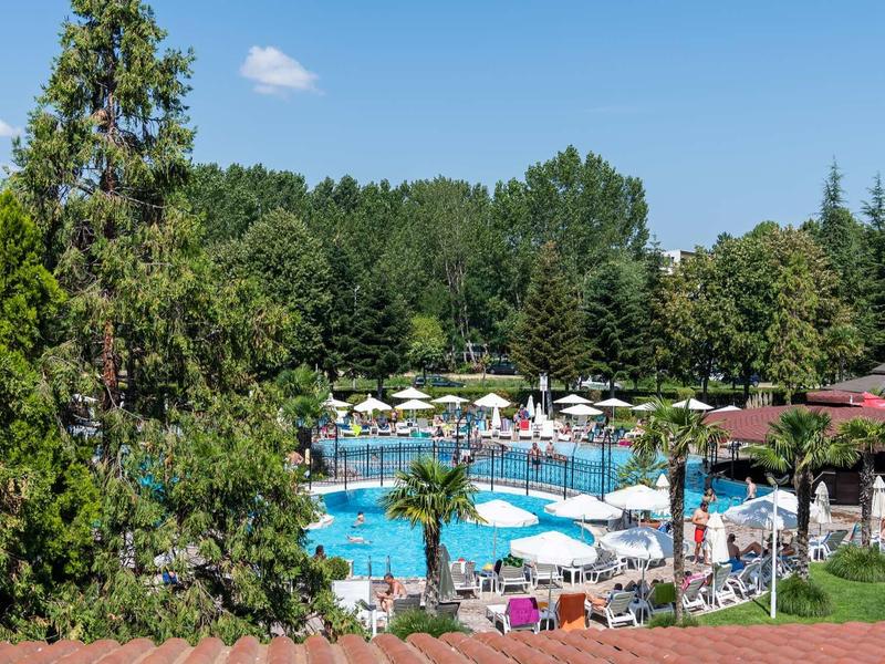 Busy pool area surrounded by trees and umbrellas in a lush hotel setting.