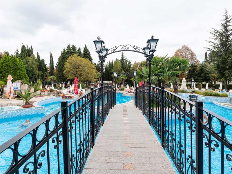 Bridge with black railings over a pool, surrounded by garden furniture and trees under a cloudy sky.