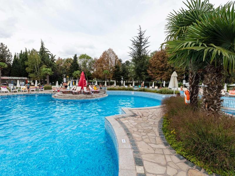 Outdoor pool with sun umbrellas and loungers surrounded by trees and palms in a hotel garden.