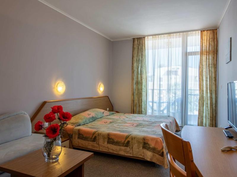 Bright hotel room with double bed, desk, TV, and balcony with sheer curtains.