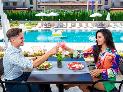 Couple sits at poolside table, clinking drinks and enjoying a meal.