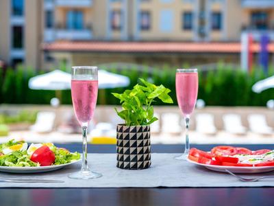 Two glasses with pink drink, plates with salad and tomatoes, small plant on table by pool