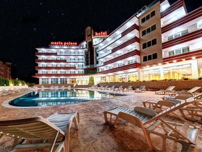 Vista nocturna de un hotel iluminado con piscina y tumbonas en primer plano.