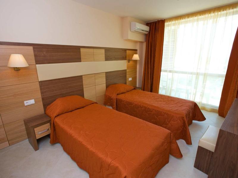 Two single beds with orange bedspreads in a bright hotel room with large window.