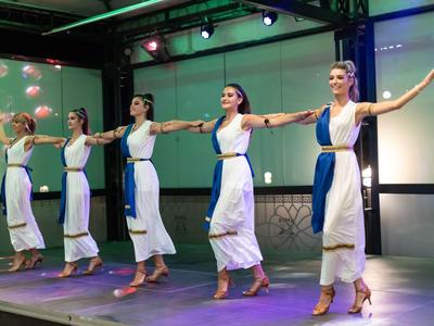 Vijf vrouwen dansen synchroon op een podium in wit-blauwe jurken.