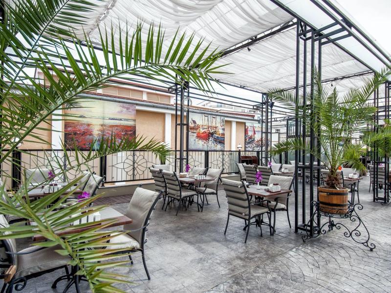 Overdekt terras met tafels, stoelen, planten en uitzicht op gebouwen.