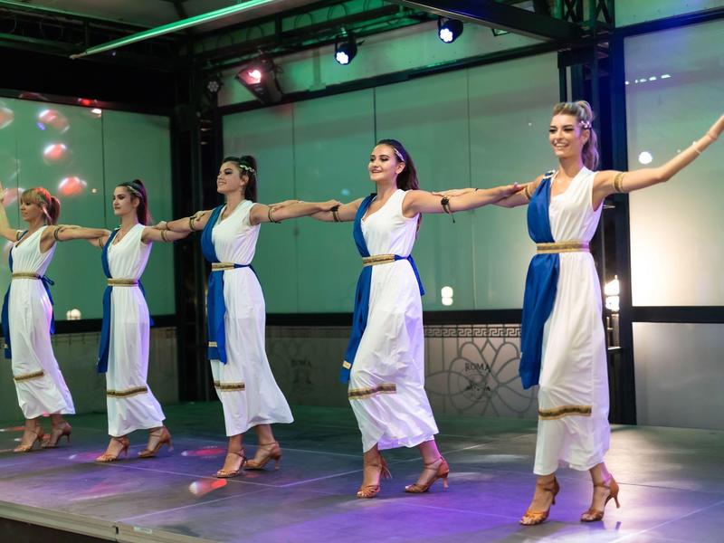 Vijf vrouwen dansen op een podium in witte jurken met blauwe sjerpen.