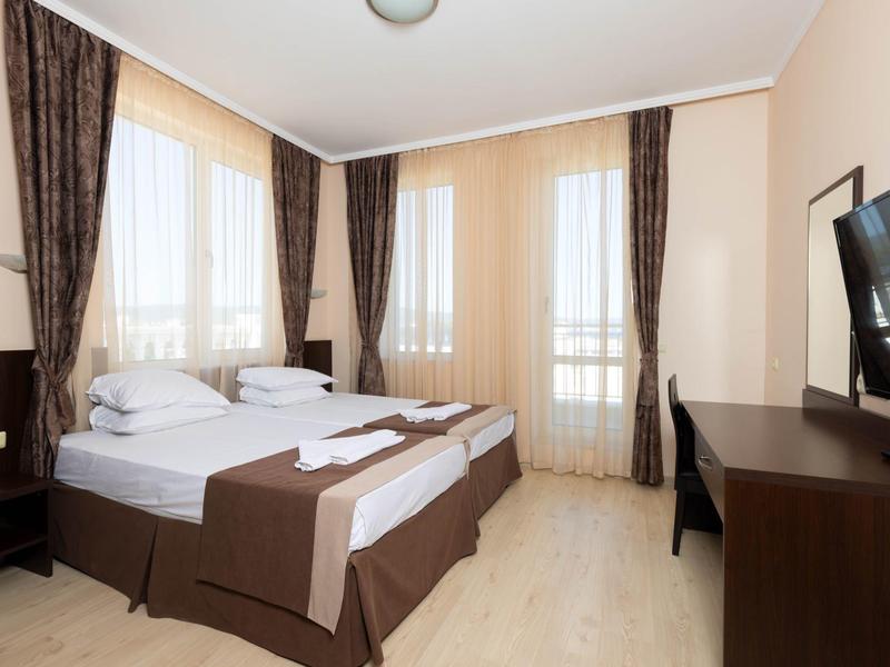 Lichte hotelkamer met twee eenpersoonsbedden, bruine gordijnen en een tv aan de muur.