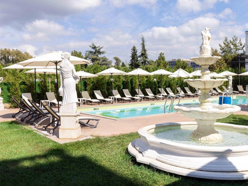 Hotelzwembad met ligstoelen, parasols en beeldhouwwerken onder een blauwe lucht.