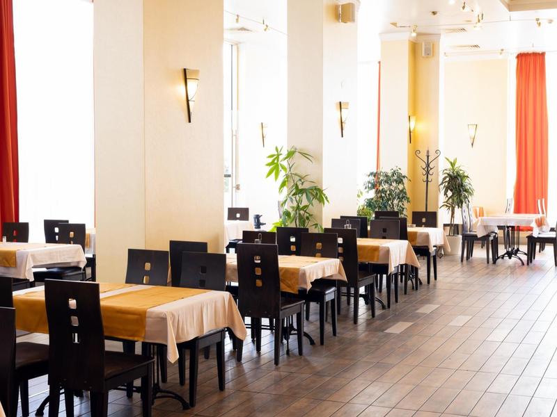 Lichte eetzaal met houten tafels, zwarte stoelen en grote ramen met rode gordijnen.