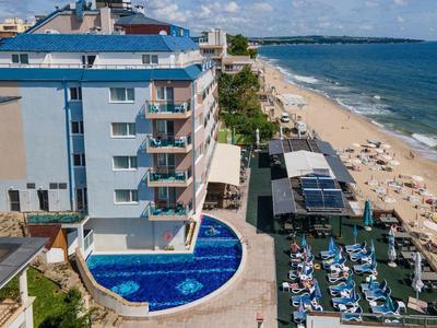 Helles Hotelgebäude mit Pool, Sonnenliegen und sandigem Strand am blauen Meer unter blauem Himmel.