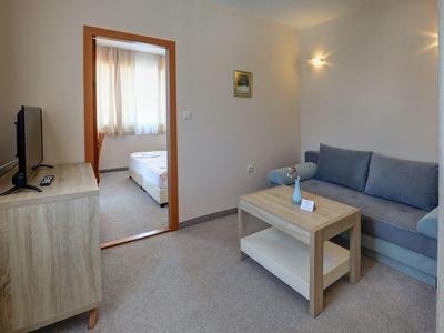 Kleines Wohnzimmer mit grauem Sofa, Holztisch und TV, daneben Blick ins Schlafzimmer mit Bett.