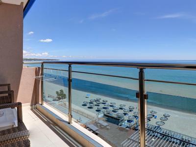 Balkon mit Glasgeländer, Korbmöbeln und Blick auf Strand, Meer und blauen Himmel.