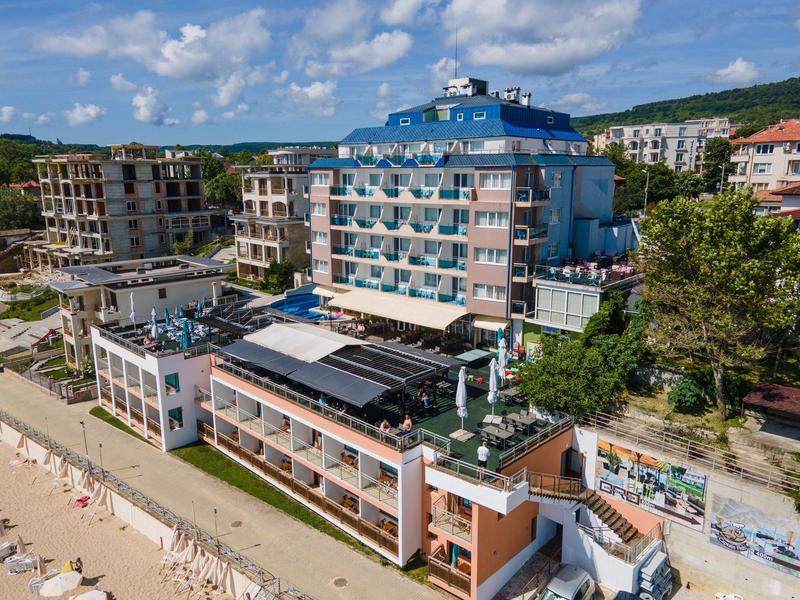 Mehrstöckiges Hotelgebäude mit Balkon, angrenzendem Strand und Meer, umgeben von grünen Hügeln.