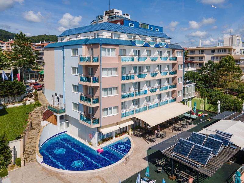Mehrstöckiges Hotel mit blauen Dächern, Swimmingpool und Terrasse, umgeben von Grünflächen.
