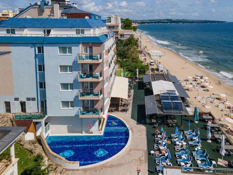 Helles Hotelgebäude mit Pool, Sonnenliegen und sandigem Strand am blauen Meer unter blauem Himmel.