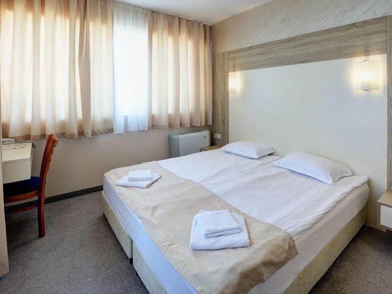 Kleines helles Schlafzimmer mit Doppelbett, weißen Bettlaken, Handtüchern und einem Schreibtisch am Fenster.