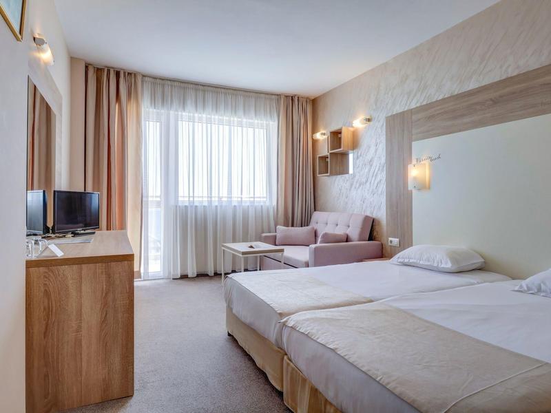 Helles Hotelzimmer mit Doppelbett, Sofa, Fernseher auf Holzmöbel und großem Fenster mit Vorhängen.