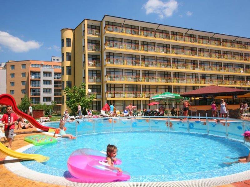 Runder Außenpool mit spielenden Kindern vor einem mehrstöckigen Hotel und blauem Himmel.