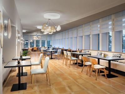 Moderner Speisesaal mit Stühlen und Tischen, großen Fenstern und stilvollen Lampen.