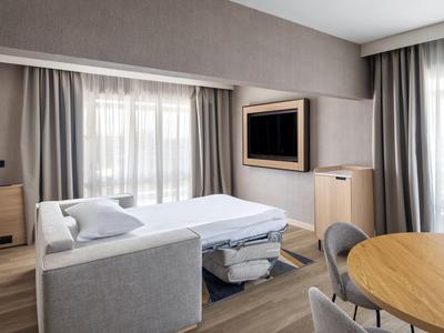 Modern ingerichte hotelkamer met bank, uitschuifbaar bed, tafel en stoelen bij ramen met gordijnen.