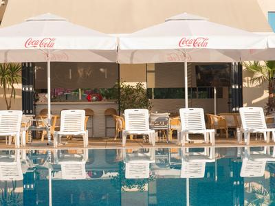 Schwimmbad mit weißen Liegestühlen und Coca-Cola-Sonnenschirmen vor einem Gebäude.