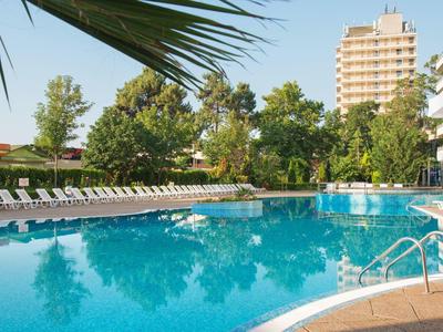 Large hotel pool with sun loungers surrounded by trees and high-rise building in the background.