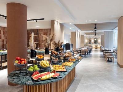 Modernes Hotelrestaurant mit Buffet, verschiedenen Früchten und Sitzbereichen in heller Atmosphäre.