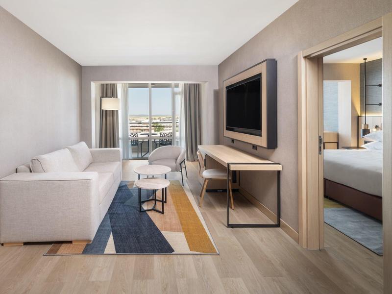 Modern hotel room with sofa, TV, rug, and view into the bedroom.