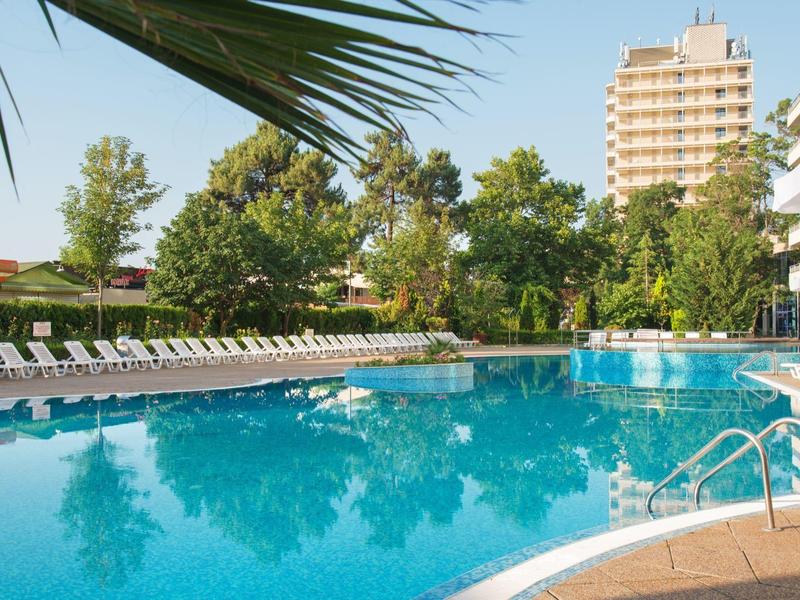 Large hotel pool with sun loungers surrounded by trees and high-rise building in the background.