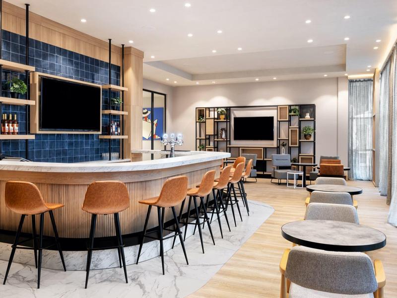 Modern hotel bar with wooden counter, bar stools, and cozy lounge area next to large windows.