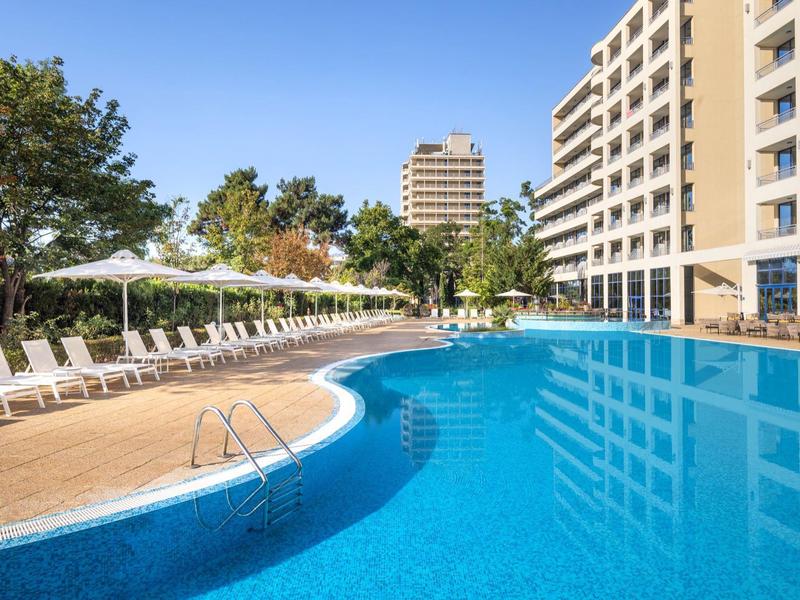 Large outdoor pool with sun loungers and hotel buildings on a sunny day.