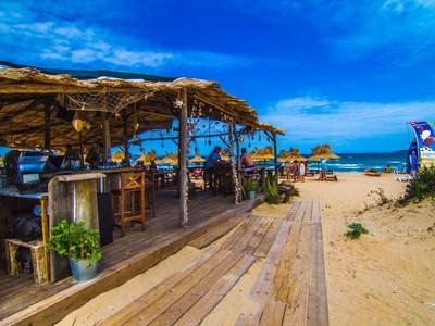 Offenes Strandcafé mit Holzdach, Tischen, Stühlen und Blick auf das blaue Meer und den Sandstrand.
