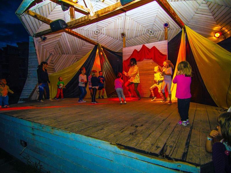 Kinder spielen auf einer beleuchteten Holz-Bühne mit bunten Vorhängen in der Nacht.