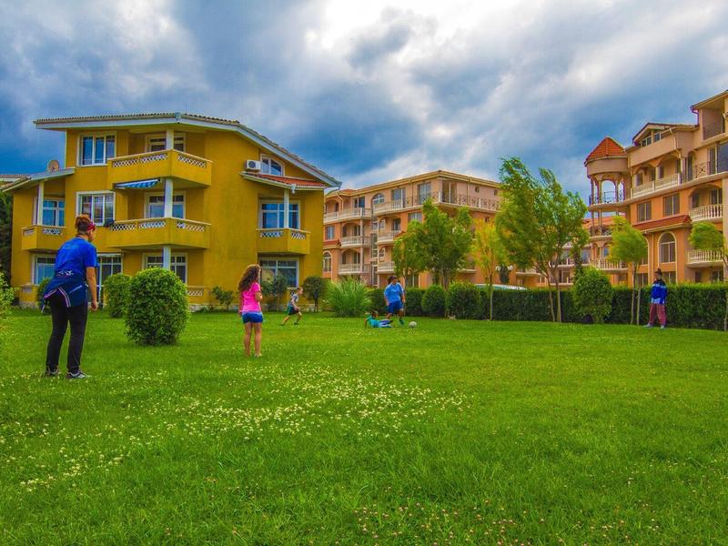 Eine Gruppe spielt Fußball auf grünem Rasen vor bunten Wohnhäusern bei bewölktem Himmel.