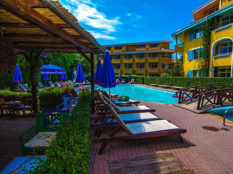 Großer Außenpool mit Liegestühlen, Sonnenschirmen und bunten Hotelgebäuden bei blauem Himmel.