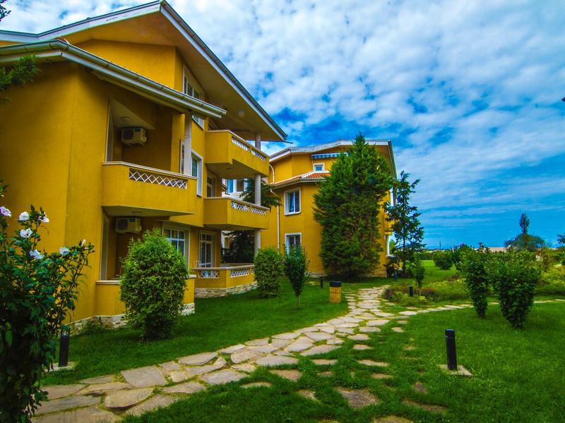Gelbes zweistöckiges Haus mit Balkon, umgeben von grünem Garten und blauem Himmel.