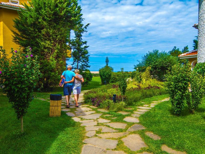 Zwei Personen gehen auf einem Steinweg zwischen grünen Gärten mit bunten Häusern bei blauem Himmel.