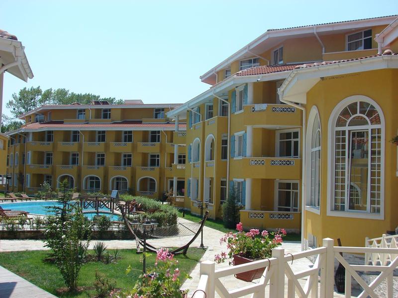 Gelbes Hotelgebäude mit Swimmingpool, grünen Pflanzen und Blumen vor blauem Himmel.