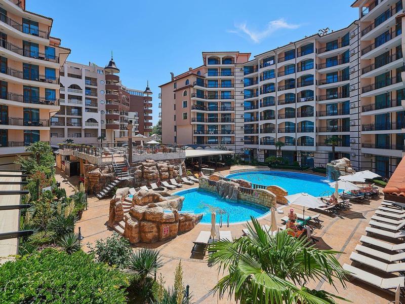 Moderner Hotelkomplex mit Schwimmbad, Liegestühlen, Palmen und blauem Himmel im Hintergrund.