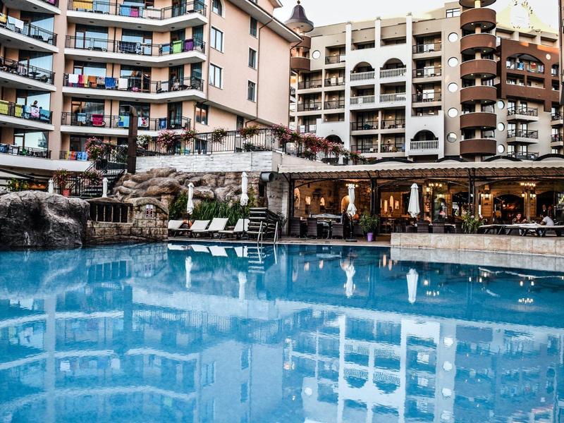 Großes Hotel mit Balkonzimmern und blauem Pool im Vordergrund, umgeben von Liegestühlen.