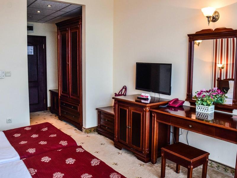 Hotelzimmer mit dunkelrotem Teppich, Holzkommode, Spiegel, Fernseher und Vase mit Blumen.