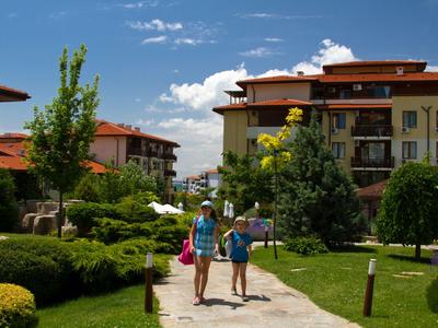 Dwie kobiety niosące torby idą ścieżką w hotelowym ogrodzie z zielonymi krzewami i kwiatami.