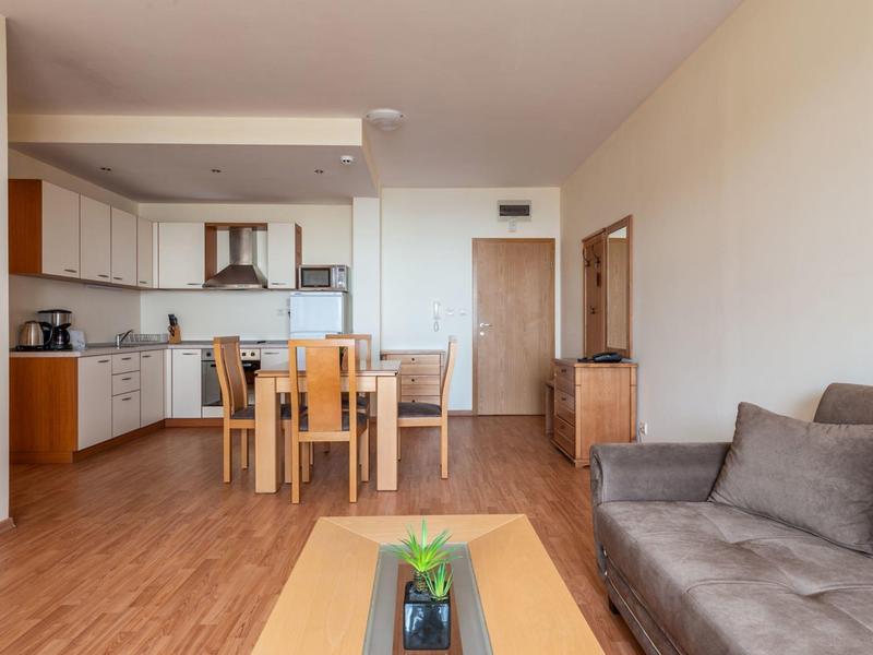 Lichte woonkamer met grijze bank, houten tafel, eettafel en moderne kitchenette.