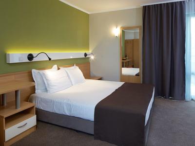 Moderne hotelkamer met tweepersoonsbed, nachtkastjes, groene muur en groot raam met gordijn.