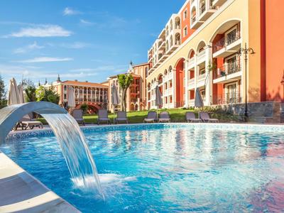 Piscina moderna dell'hotel con cascata sotto il sole e edificio a più piani sullo sfondo.