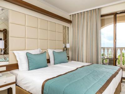 Hell eingerichtetes Schlafzimmer mit Doppelbett, türkisfarbenen Kissen und Decke, Balkon und Vorhängen.